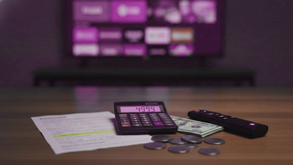 A calculator, dollar bills, coins, a streaming stick and a remote on a table in front of a TV