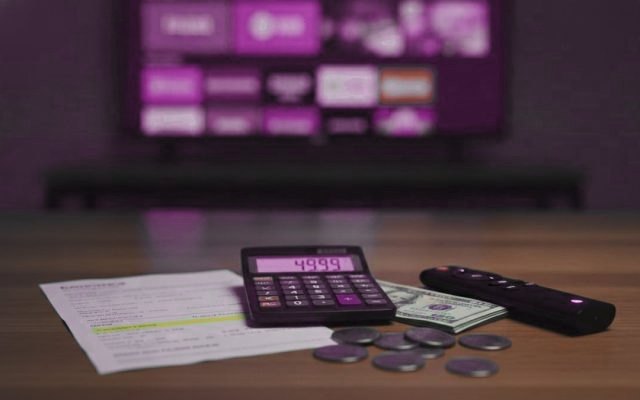 A calculator, dollar bills, coins, a streaming stick and a remote on a table in front of a TV